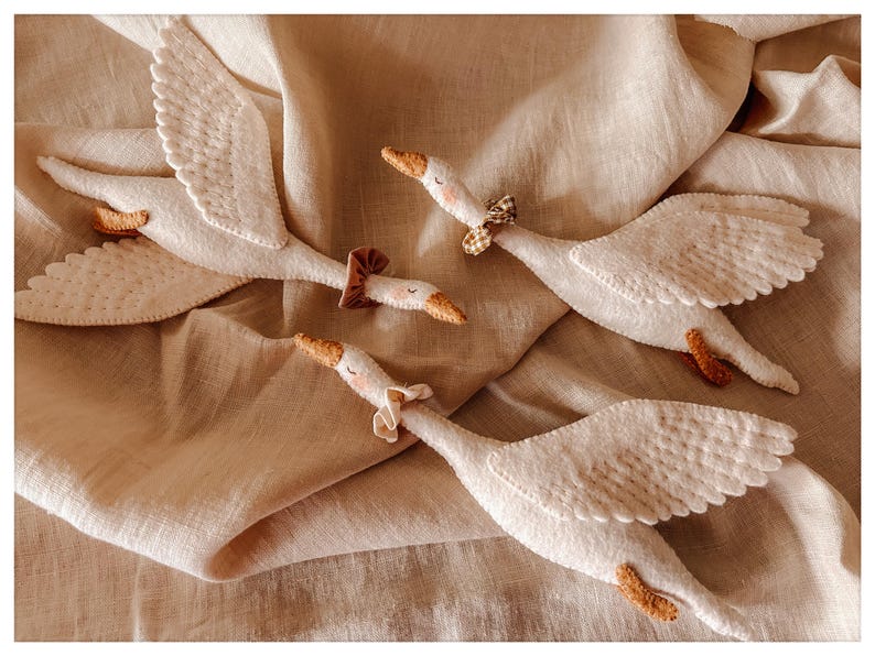 May include: Four white felt goose toys with outstretched wings and small, decorative bows around their necks. The geese have orange beaks and feet, and are arranged on a beige fabric background.