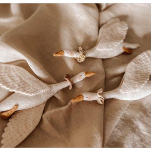 May include: Four white felt goose toys with outstretched wings are arranged on a beige fabric surface. Each goose has a tan beak and feet, with a decorative collar around its neck. The toys appear to be handmade.