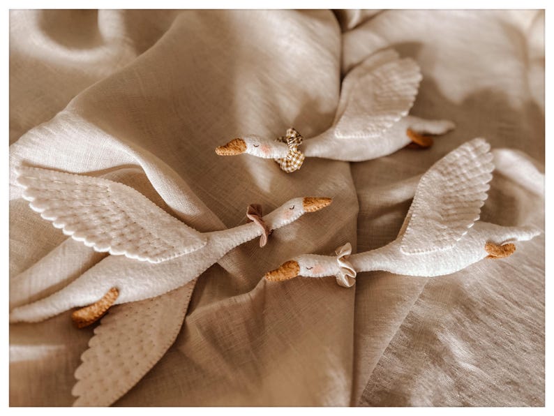 May include: Four white felt goose toys with outstretched wings are arranged on a beige fabric surface. Each goose has a tan beak and feet, with a decorative collar around its neck. The toys appear to be handmade.