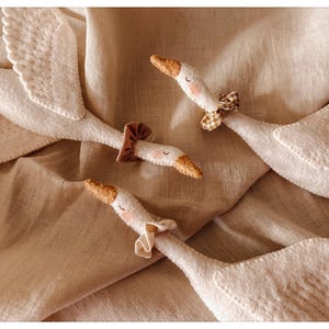 May include: A collection of white felt geese with detailed wings and orange beaks. Each goose has a decorative neck accessory, including a brown mushroom cap and patterned bows. The geese are arranged on a tan fabric background.