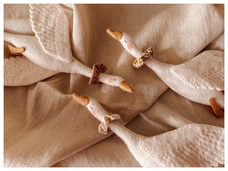 May include: A collection of white felt geese with detailed wings and orange beaks. Each goose has a decorative neck accessory, including a brown mushroom cap and patterned bows. The geese are arranged on a tan fabric background.