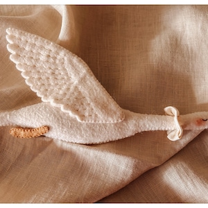 May include: A handmade white felt goose toy with outstretched wings, a tan beak, and small tan feet. The goose has a small bow around its neck and is set against a neutral linen background.