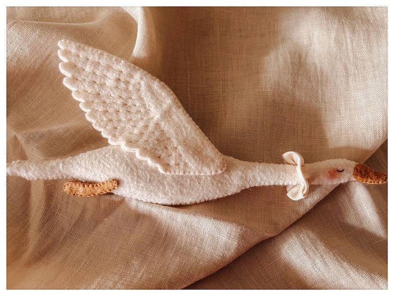 May include: A handmade white felt goose toy with outstretched wings, a tan beak, and small tan feet. The goose has a small bow around its neck and is set against a neutral linen background.