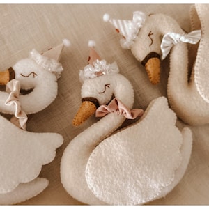 May include: Three plush swan toys with white bodies, brown beaks, and closed eyes. Each swan wears a party hat and a bow or ruffle around its neck. The swans are arranged on a neutral-toned fabric.