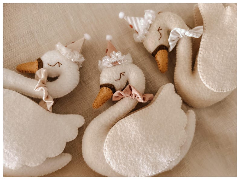 May include: Three plush swan toys with white bodies, brown beaks, and closed eyes. Each swan wears a party hat and a bow or ruffle around its neck. The swans are arranged on a neutral-toned fabric.