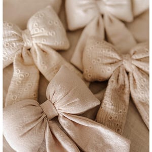 Puede incluir: Una colección de lazos de tela beige, algunos con detalles de calado. Los lazos están dispuestos sobre una superficie de tonos neutros, creando una estética suave y delicada. Los lazos varían en tamaño y textura, ofreciendo una gama de opciones para accesorios para el cabello o proyectos de manualidades.