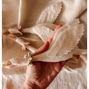 May include: Handmade white felt swan toys with detailed wings, yellow beaks, and small pink cheeks. Each swan has a decorative bow. The toys are displayed on a cream-colored fabric, perfect for nursery decor or children's play.