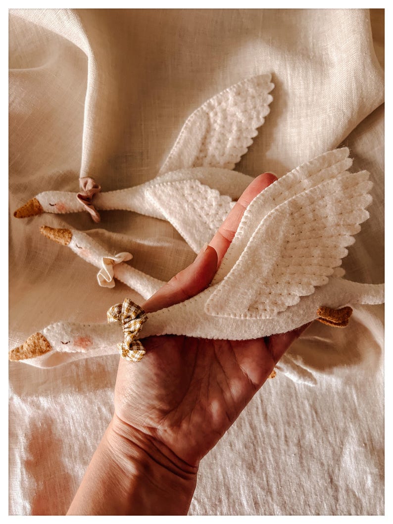 May include: Handmade white felt swan toys with detailed wings, yellow beaks, and small pink cheeks. Each swan has a decorative bow. The toys are displayed on a cream-colored fabric, perfect for nursery decor or children's play.