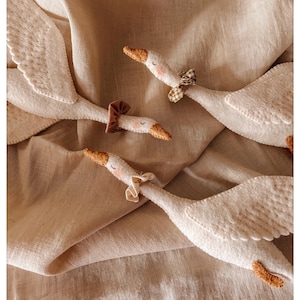 May include: Close-up of several white felt goose toys with brown beaks and small bows around their necks. The geese are arranged on a beige linen fabric, creating a soft and whimsical scene. The wings are detailed with stitched patterns.