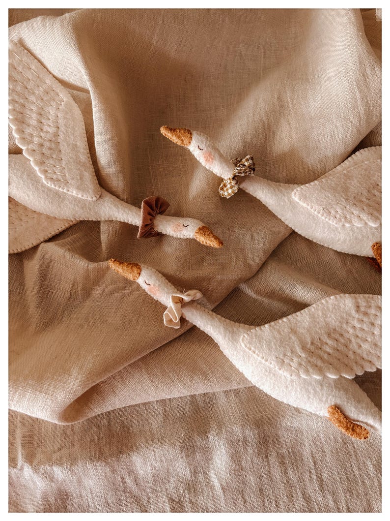 May include: Close-up of several white felt goose toys with brown beaks and small bows around their necks. The geese are arranged on a beige linen fabric, creating a soft and whimsical scene. The wings are detailed with stitched patterns.