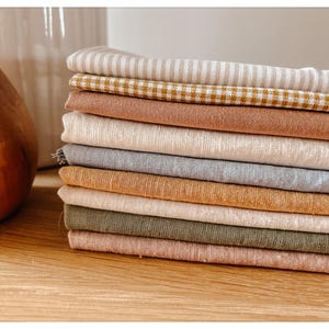 May include: A stack of folded fabric in various colors, including beige, brown, and green. Some pieces have striped or checkered patterns. The fabrics are arranged on a wooden surface, suggesting a textile or craft theme.