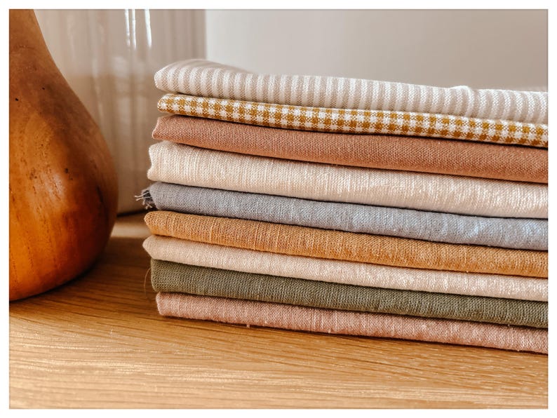 May include: A stack of folded fabric in various colors, including beige, brown, and green. Some pieces have striped or checkered patterns. The fabrics are arranged on a wooden surface, suggesting a textile or craft theme.