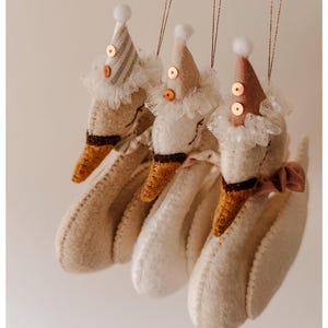 May include: Three handmade goose ornaments with party hats and lace collars. The geese are white with brown beaks and feet. Each goose wears a different colored party hat with sequins and a small white pom-pom on top. A brown ribbon bow adorns one goose.