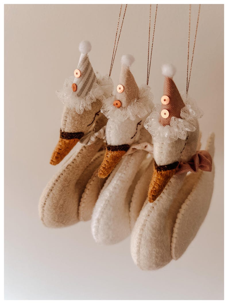 May include: Three handmade goose ornaments with party hats and lace collars. The geese are white with brown beaks and feet. Each goose wears a different colored party hat with sequins and a small white pom-pom on top. A brown ribbon bow adorns one goose.