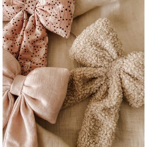 Può includere: Tre fiocchi di tessuto in diverse trame e colori. Un fiocco è rosa chiaro con un motivo a foglie bianche e nere. Un altro è rosa chiaro tinta unita e il terzo è un fiocco beige strutturato. I fiocchi sono disposti su un tessuto dai toni neutri.