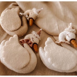 May include: A collection of handmade felt swan ornaments. The swans are primarily white with beige accents, featuring orange beaks and decorative hats. Some swans have pink bows. The ornaments are arranged on a beige fabric background.