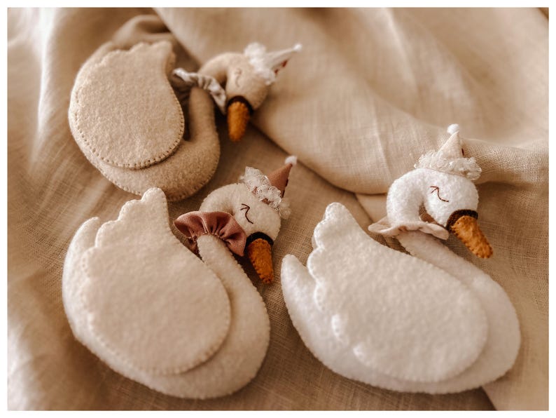 May include: A collection of handmade felt swan ornaments. The swans are primarily white with beige accents, featuring orange beaks and decorative hats. Some swans have pink bows. The ornaments are arranged on a beige fabric background.