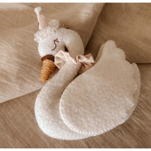 May include: A plush white swan toy with a brown beak and a small crown. The swan has a beige bow around its neck and two large, rounded wings. The toy is set against a neutral, textured background.