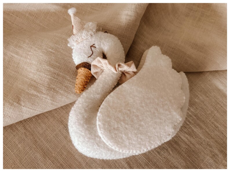 May include: A plush white swan toy with a brown beak and a small crown. The swan has a beige bow around its neck and two large, rounded wings. The toy is set against a neutral, textured background.