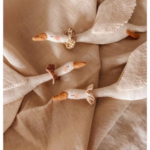 May include: Soft, white felt geese toys with outstretched wings and brown beaks. Each goose has a small bow tied around its neck. The geese are arranged on a beige fabric background, creating a whimsical and gentle scene.