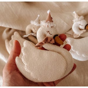 May include: Handmade felt swan toys in cream and white, adorned with small hats and bows. The swans have red beaks and are held in a hand, with a soft, neutral background. These plush toys are suitable for children.