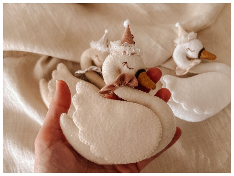 May include: Handmade felt swan toys in cream and white, adorned with small hats and bows. The swans have red beaks and are held in a hand, with a soft, neutral background. These plush toys are suitable for children.