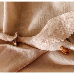 May include: A handmade white felt goose toy with a brown beak and feet, and a small patterned bow around its neck. The wings are detailed with stitched patterns. The toy is lying on a beige linen fabric.
