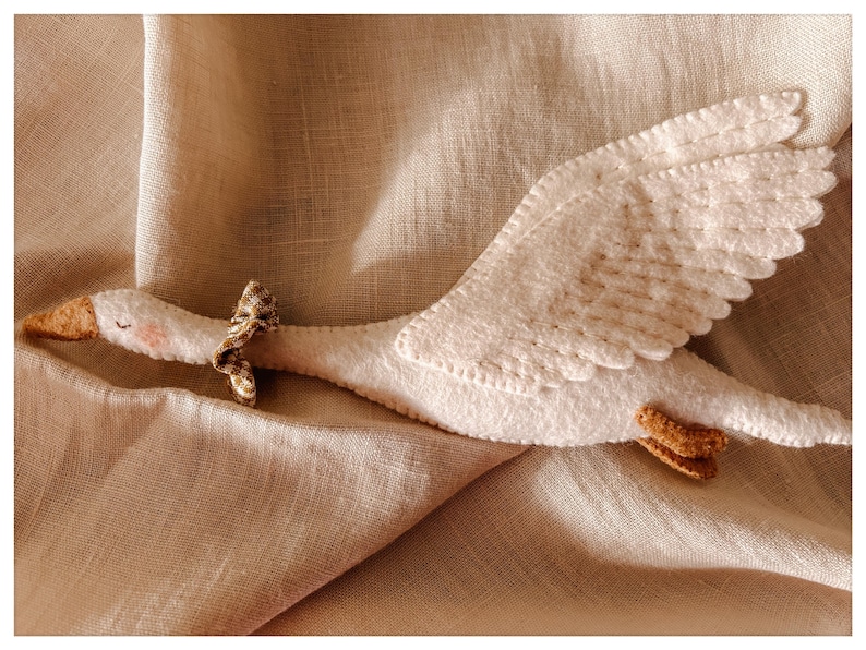 May include: A handmade white felt goose toy with a brown beak and feet, and a small patterned bow around its neck. The wings are detailed with stitched patterns. The toy is lying on a beige linen fabric.