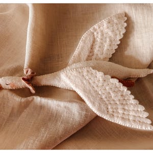 May include: A handmade white goose plush toy with detailed wings and a small pink bow. The goose has a yellow beak and is lying on a beige fabric surface. The wings are textured with a quilted pattern.