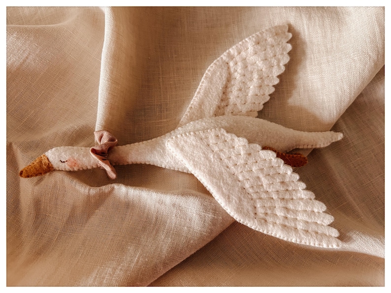 May include: A handmade white goose plush toy with detailed wings and a small pink bow. The goose has a yellow beak and is lying on a beige fabric surface. The wings are textured with a quilted pattern.