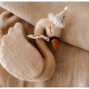 May include: A handmade, beige felt swan toy with a small white party hat and a ruffled collar. The swan has a brown beak and stitched details. The toy is placed on a beige linen fabric.