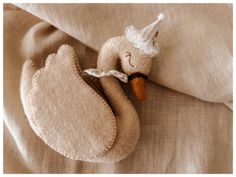 May include: A handmade, beige felt swan toy with a small white party hat and a ruffled collar. The swan has a brown beak and stitched details. The toy is placed on a beige linen fabric.