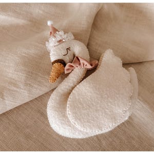May include: A plush white swan toy with a pink bow, a brown beak, and a small party hat. The swan is made of soft, textured material and has stitched details. The toy is lying on a beige fabric surface.