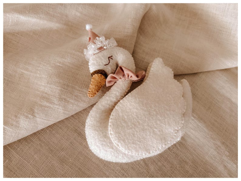 May include: A plush white swan toy with a pink bow, a brown beak, and a small party hat. The swan is made of soft, textured material and has stitched details. The toy is lying on a beige fabric surface.