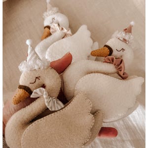 May include: Handmade felt swan ornaments in cream and beige, adorned with party hats and bows. The swans have brown beaks and stitched details, creating a whimsical and festive aesthetic. Perfect for nursery decor or a child's room.