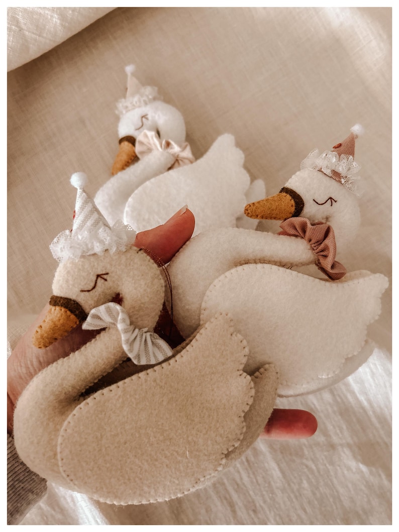 May include: Handmade felt swan ornaments in cream and beige, adorned with party hats and bows. The swans have brown beaks and stitched details, creating a whimsical and festive aesthetic. Perfect for nursery decor or a child's room.