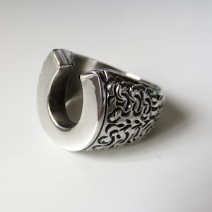 May include: A silver ring with a horseshoe design. The horseshoe is smooth and polished, while the band of the ring is textured with a floral pattern.