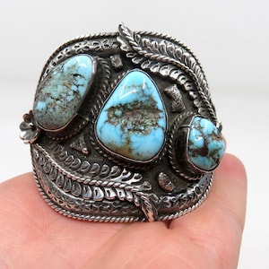 May include: A silver cuff bracelet with three turquoise stones set in a detailed design. The bracelet has a textured surface and a rope-like border.