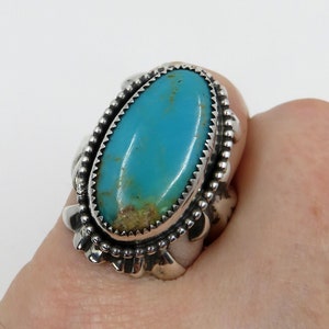 Puede incluir: Un anillo de plata con una gran piedra turquesa ovalada. La piedra es de color azul verdoso brillante con inclusiones marrones. El anillo tiene un bisel festoneado detallado y una banda decorativa.