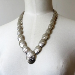 May include: A silver necklace with a large, oval pendant featuring a floral design. The necklace is made of multiple oval-shaped links.