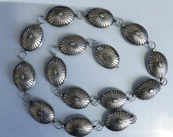 May include: A silver chain belt with oval-shaped links featuring a sunburst design. Each link is connected by small silver rings.