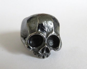 May include: A silver skull ring with a detailed design. The ring is made of metal and has a smooth, polished finish.