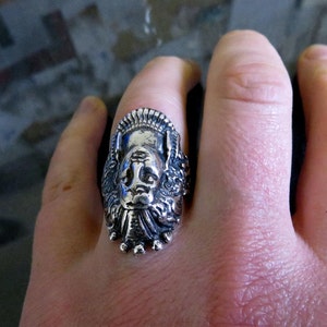 Peut inclure: Une bague en argent avec un design d&eacute;taill&eacute; d'un visage avec une couronne. La bague est port&eacute;e au doigt d'une personne.