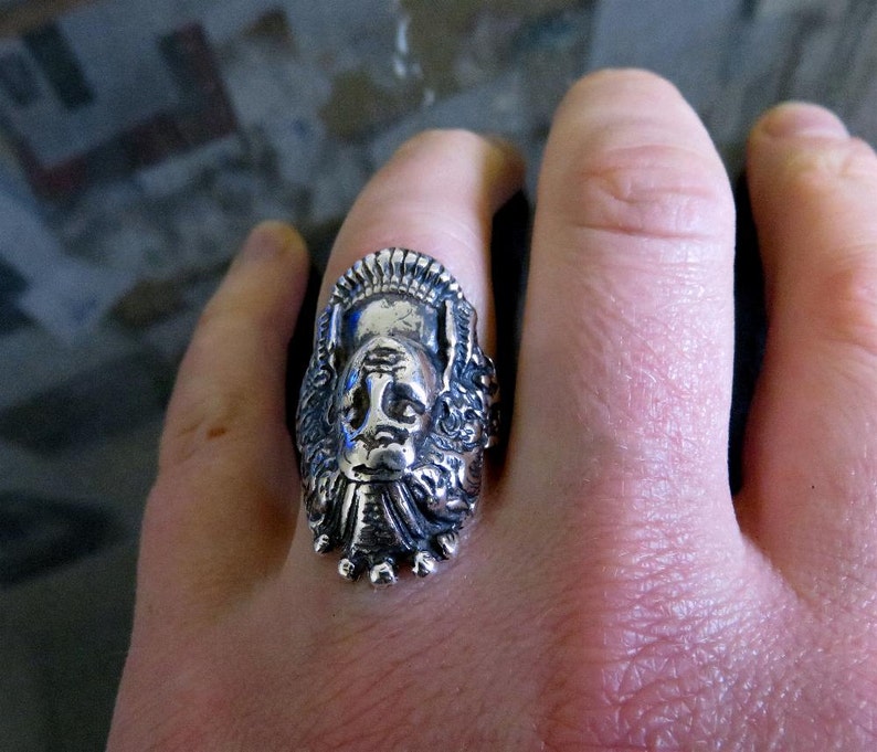 Peut inclure: Une bague en argent avec un design d&eacute;taill&eacute; d'un visage avec une couronne. La bague est port&eacute;e au doigt d'une personne.