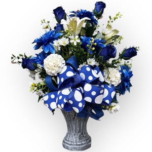 May include: A blue and white floral arrangement in a gray ceramic vase. The flowers include blue roses, white daisies, and blue and white lilies. The arrangement is tied with a blue and white polka dot bow.