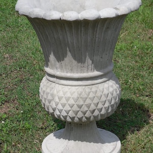May include: A large, gray concrete planter with a fluted design and a diamond-shaped pattern on the bottom. The planter is sitting on a bed of green grass.