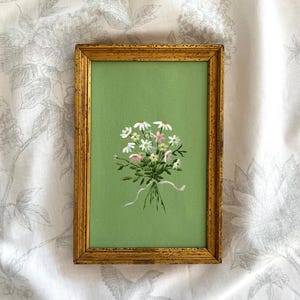 Puede incluir: Una pintura enmarcada de un ramo de margaritas blancas y rosas con hojas verdes, atado con una cinta blanca, sobre un fondo verde.