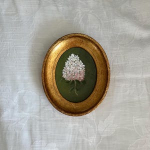 Puede incluir: Una pieza de arte mural de forma ovalada que presenta una pintura detallada de un racimo de flores blancas y rosa claro sobre un fondo verde oscuro, encerrada en un marco dorado texturizado y ornamentado con un borde interior perlado.