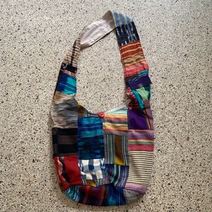 Peut inclure: Un sac à bandoulière patchwork avec une longue sangle. Le sac est composé de carrés de tissu de différentes couleurs, notamment rouge, bleu, violet et marron. Un bouton rouge ferme l'ouverture du sac. Le sac est conçu pour transporter des objets.