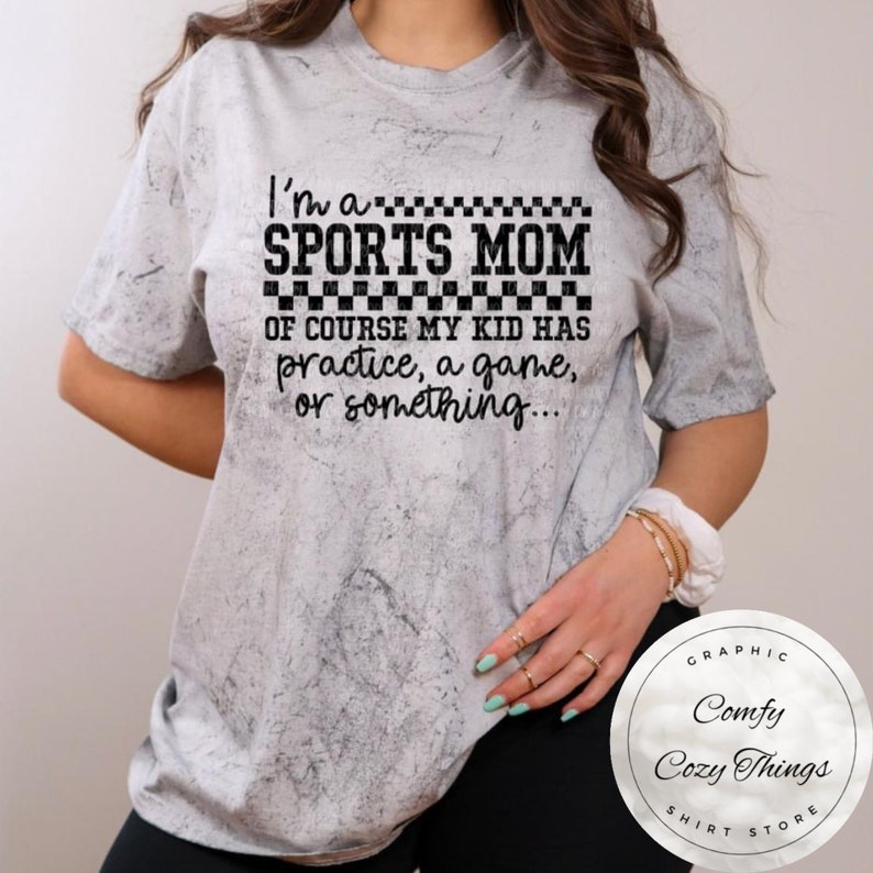 May include: A white t-shirt with a black and white checkered design that says "I'm a SPORTS MOM OF COURSE MY KID HAS practice, a game, or something..." The shirt is on a model with a white background.
